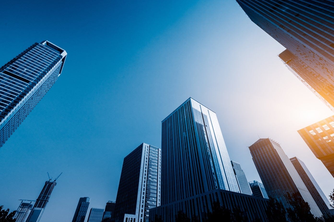 Looking up at tall office buildings with blue sky and sun visible on right side.
